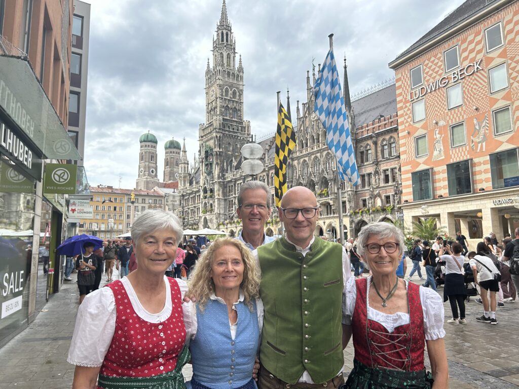 Das Foto zeigt eine Gruppe von fünf Personen (drei Frauen und 2 Männer) vor dem Neuen Rathaus in München, Deutschland. Sie tragen traditionelle bayerische Tracht. Im Hintergrund ist die gotische Architektur des Neuen Rathauses zu sehen, mit seiner aufwendigen Fassade und dem hohen Turm. Der Platz ist belebt mit Menschen und mit bayerischen Fahnen geschmückt.