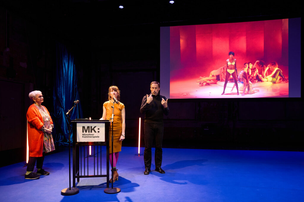 Das Bild zeigt eine Pressekonferenz der Münchner Kammerspiele zur Spielzeit 2024/25. Links steht eine ältere Frau in einem orangefarbenen Mantel, in der Mitte eine Frau an einem Rednerpult mit dem Logo der Münchner Kammerspiele, und rechts daneben ein Mann, der alles in Gebärdensprache übersetzt. Im Hintergrund ist eine Projektion mit einer Bühnenperformance, bei der eine Person in Unterwäsche im Vordergrund steht, während andere Personen im Hintergrund liegen