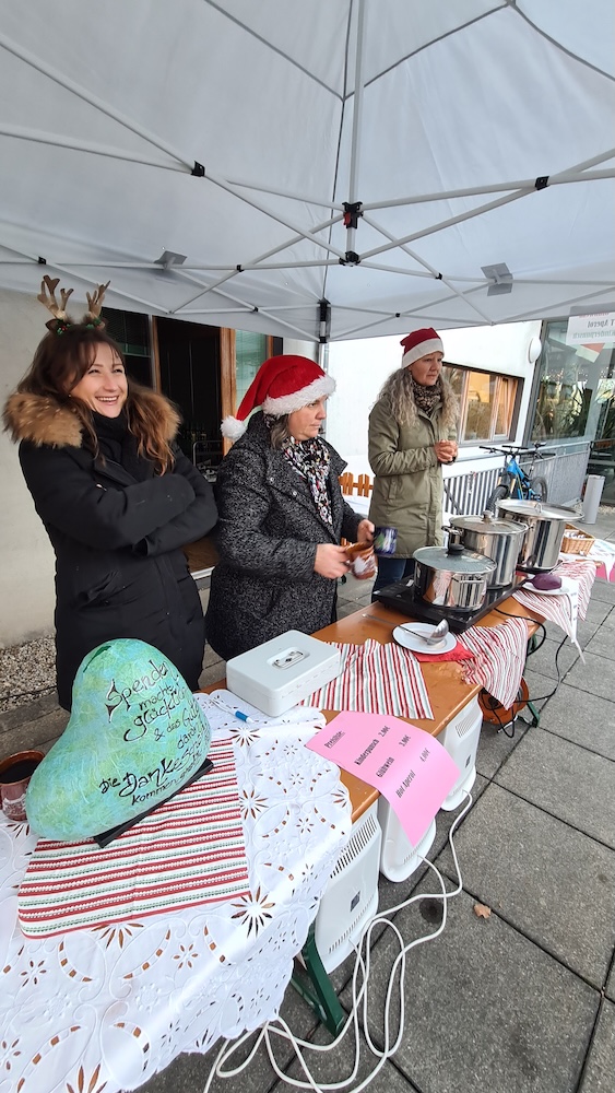 Unter einem Pavillon stehen drei Frauen, zwei haben eine Nikolausmütze. 
Davor sind festlich geschmückte Tische mit 3 Kochtöpfen.