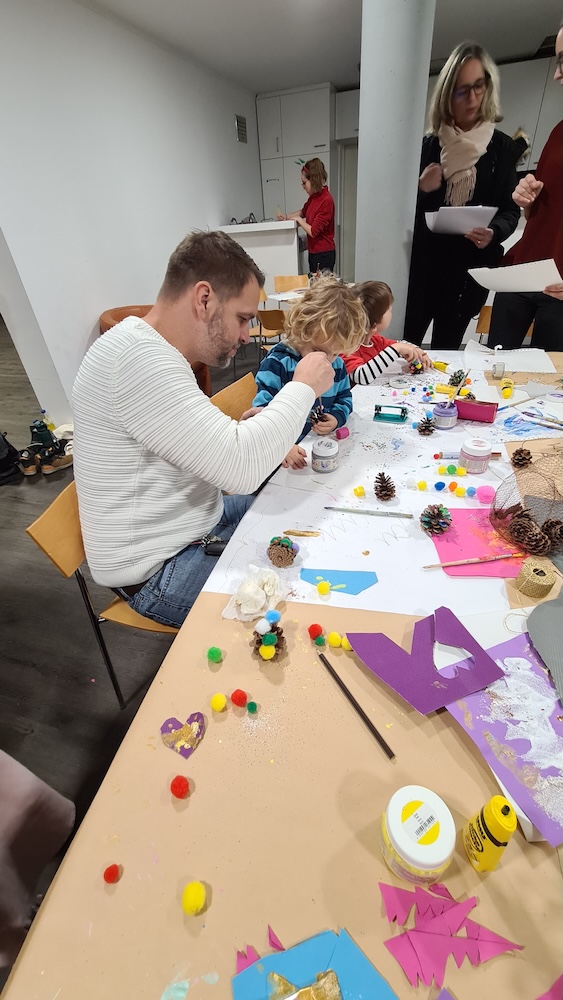 Ein Tisch mit vielen Bastelutensilien, ein Mann sitzt am Tisch und bastelt etwas, hinter ihm sitzen zwei Kinder, die auch basteln. 