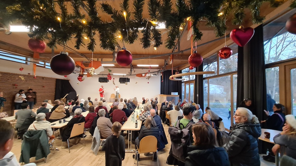 eine festlich dekorierte Veranstaltung mit weihnachtlichem Thema. Es sind viele Menschen an langen Tischen in einem Raum mit großen Fenstern versammelt. Im Vordergrund hängen grüne Girlanden mit roten Kugeln und Herzen von der Decke. Im Hintergrund findet auf einer kleinen Bühne eine Aufführung statt, die von einer Person im Weihnachtsmannkostüm moderiert wird